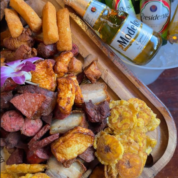 Dominican platter with fried meats, plantains, and yuca, garnished with an orchid, alongside beers.