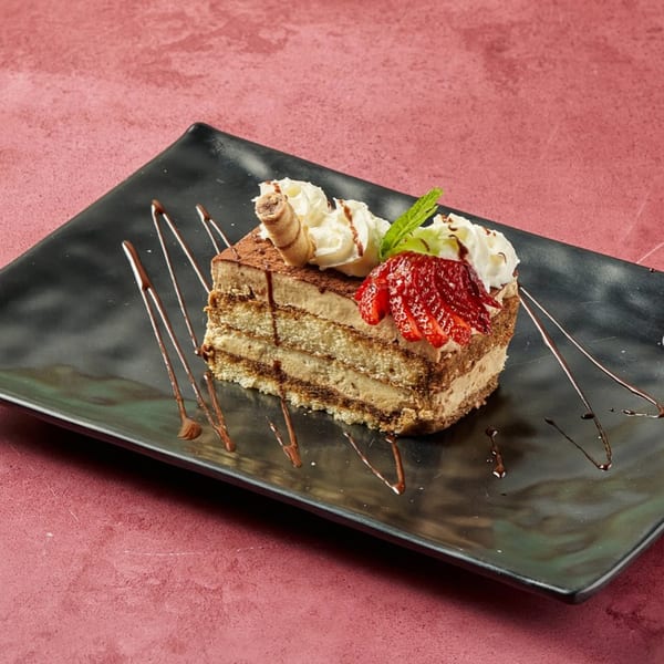 Layered tiramisu cake with whipped cream, strawberry slices, and chocolate drizzle on a black plate.