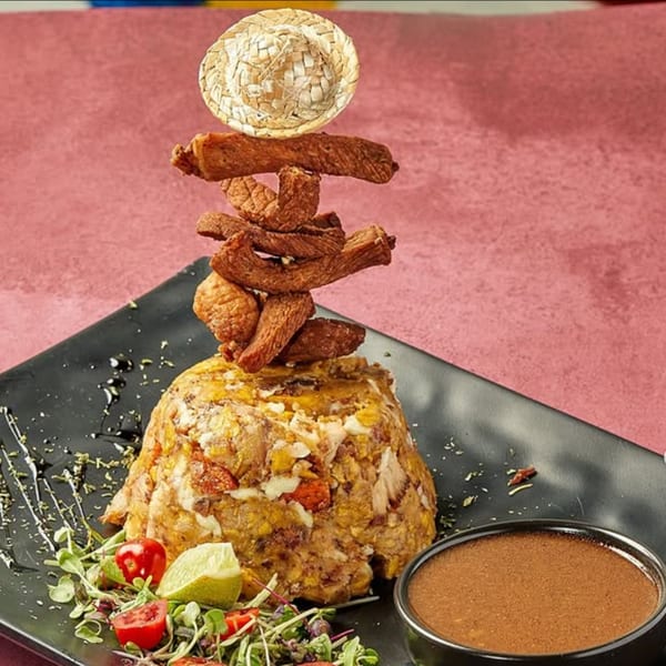 Dominican mofongo with fried meat, a small straw hat, salad, and sauce on a black plate.