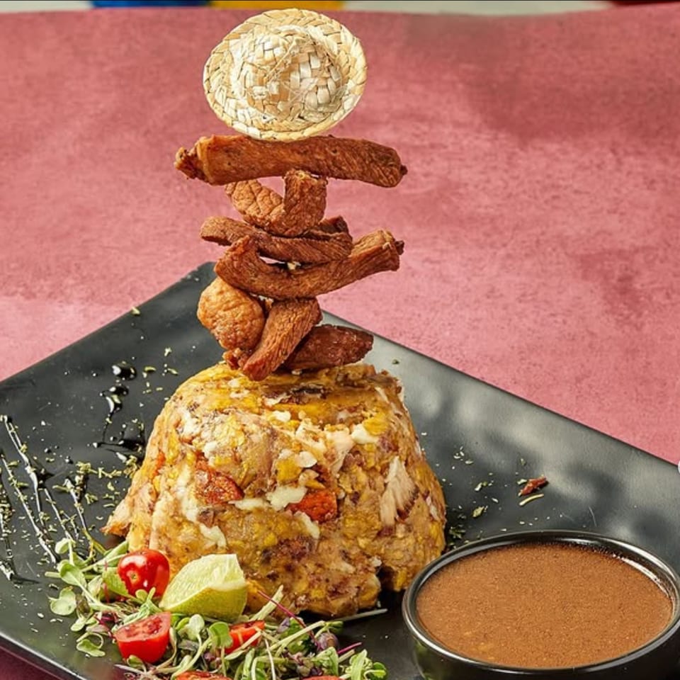 Dominican mofongo with fried meat, a small straw hat, salad, and sauce on a black plate.