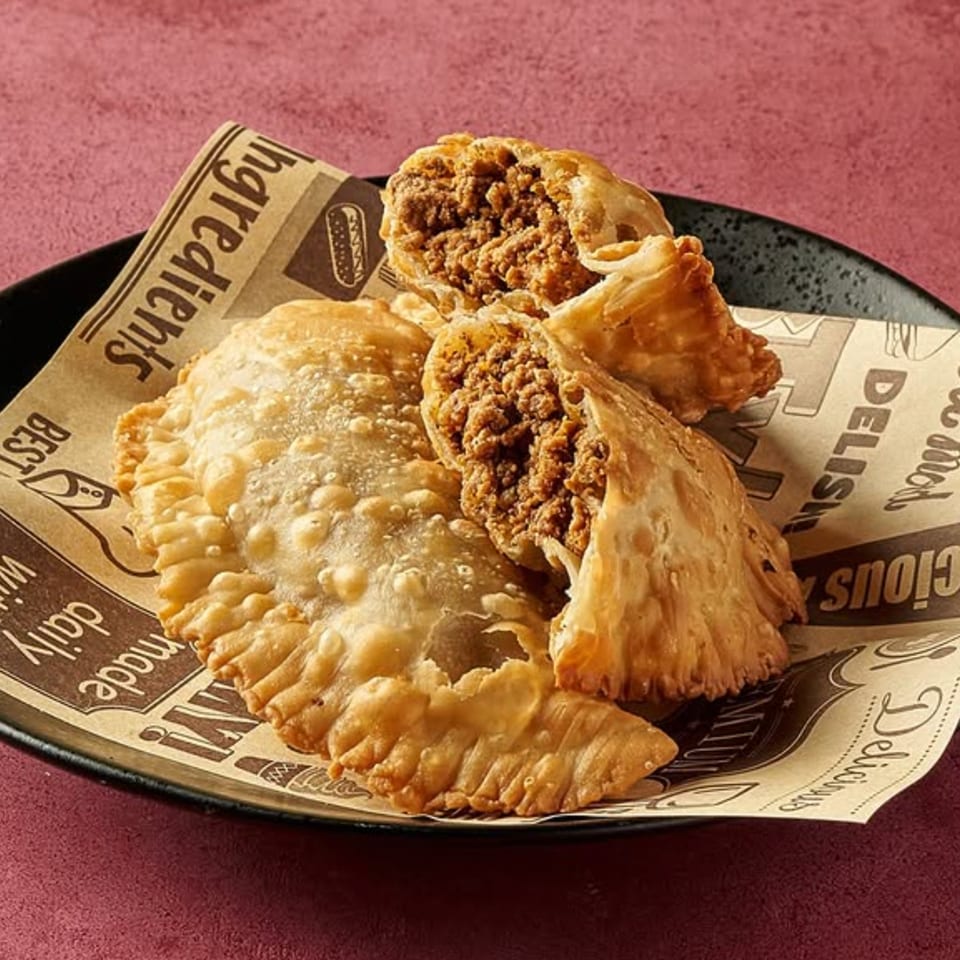 Three golden-brown Dominican empanadas, one broken open to reveal a savory meat filling, on a black plate.
