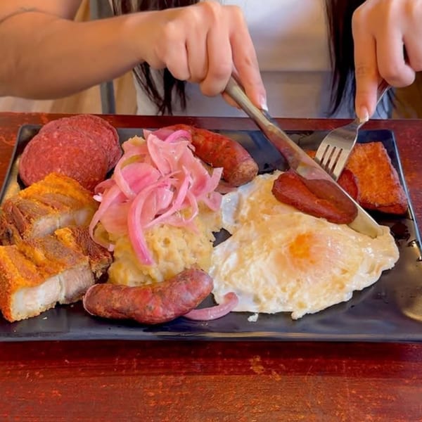 Hands cutting into a large Dominican breakfast platter with eggs, sausage, fried pork, and pickled onions.