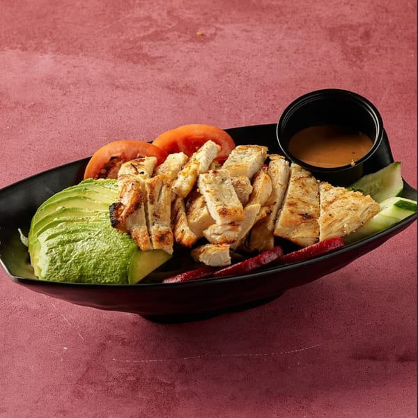 Grilled chicken salad with avocado, tomatoes, cucumbers, and a side of dressing in a black bowl.